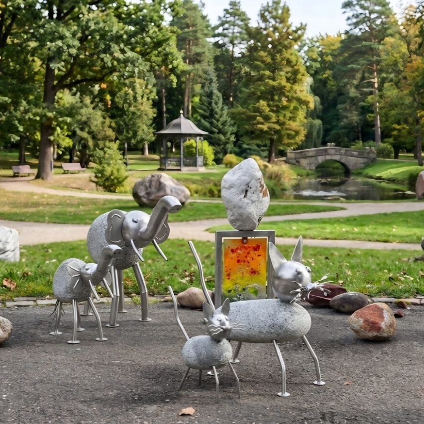 Metalltierskulpturen in einem Park, umgeben von Bäumen, einem Teich und einer Brücke. Ein Pavillon und Bänke sind im Hintergrund zu sehen.