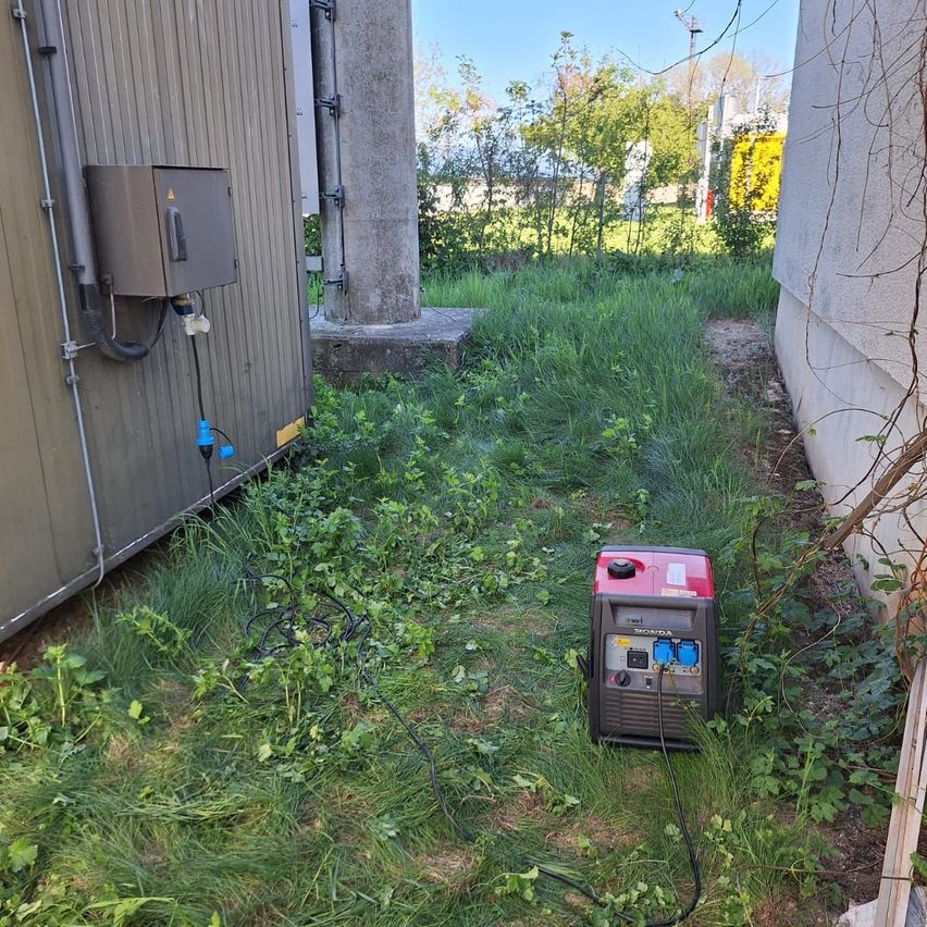 Ein roter und schwarzer tragbarer Generator steht in einem Grasbereich neben einem Gebäude, mit überwucherten Unkräutern und einem Betonpfeiler in der Nähe.