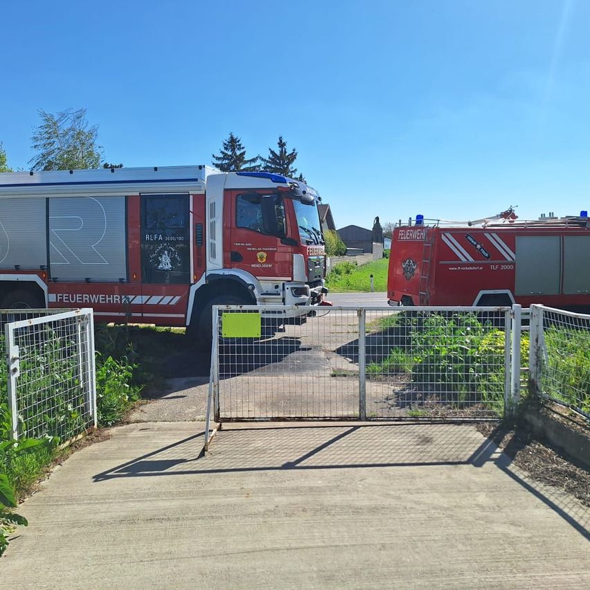Zwei Feuerwehrfahrzeuge sind in der Nähe eines geschlossenen Tors mit einem gelben Schild geparkt. Die Fahrzeuge sind rot und haben Notfallleuchten oben auf.
