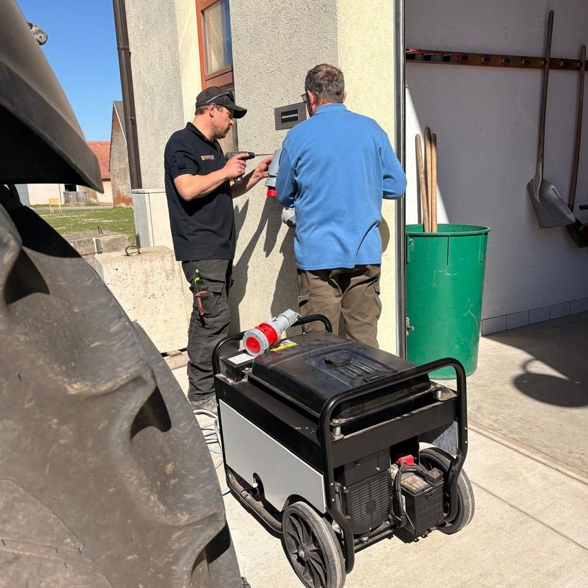 Zwei Männer arbeiten an einem Generator vor einem Gebäude. Einer bohrt in eine Wand, während der andere daneben steht. Ein großer Traktorreifen ist sichtbar.