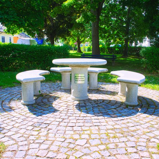 Ein Steintisch und Bänke sind in einem Park im Kreis angeordnet. Dahinter befindet sich eine Bank und eine Hecke. Im Hintergrund stehen Bäume und Häuser.