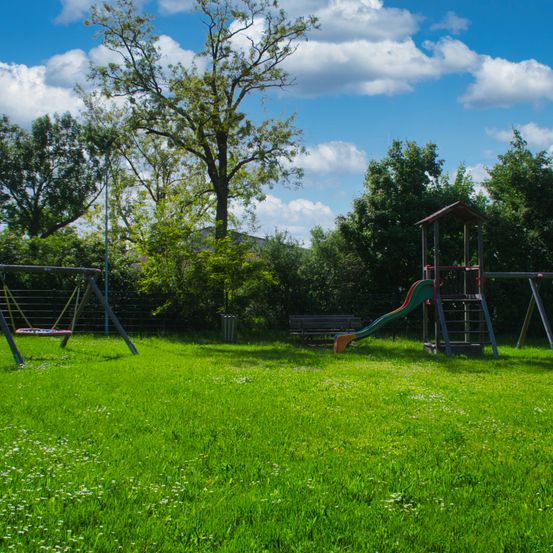 Ein Kinderspielplatz in einem Park mit einer Rutsche, Schaukeln und einer Bank unter einem blauen Himmel mit Wolken.