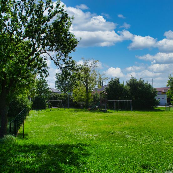 Ein sonniger Park mit grünem Rasen und Bäumen, ein Spielplatz mit Schaukeln und einer Rutsche. Der Himmel ist blau mit weißen Wolken. In der Ferne Häuser und eine Straßenlaterne.