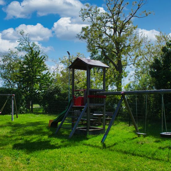 Ein Spielplatz in einem Park mit Rutsche, Schaukeln und einem Reifenschaukel unter einem blauen Himmel mit verstreuten Wolken. Grüner Rasen und Bäume umgeben das Spielareal.