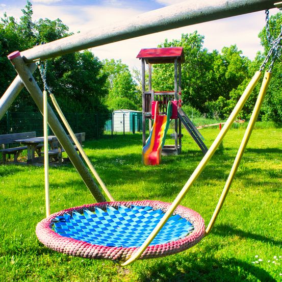 Ein bunter Spielplatz mit Schaukel und Rutsche. Die Schaukel hat einen bunten Sitz und die Rutsche ist mehrfarbig. Bäume und eine Bank sind im Hintergrund.