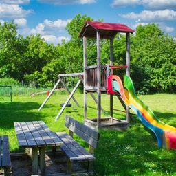 Ein Spielplatz mit einer Rutsche, einer Schaukel und einer Bank unter einem sonnigen Himmel mit grünen Bäumen im Hintergrund.