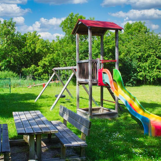 Ein Spielplatz mit einer Rutsche, einer Schaukel und einer Bank unter einem sonnigen Himmel mit grünen Bäumen im Hintergrund.