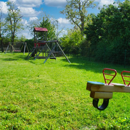 Ein Spielplatz im Freien mit Schaukeln, einer Rutsche und einem Wippstuhl an einem sonnigen Tag in einer Rasenfläche, umgeben von Bäumen und Büschen.