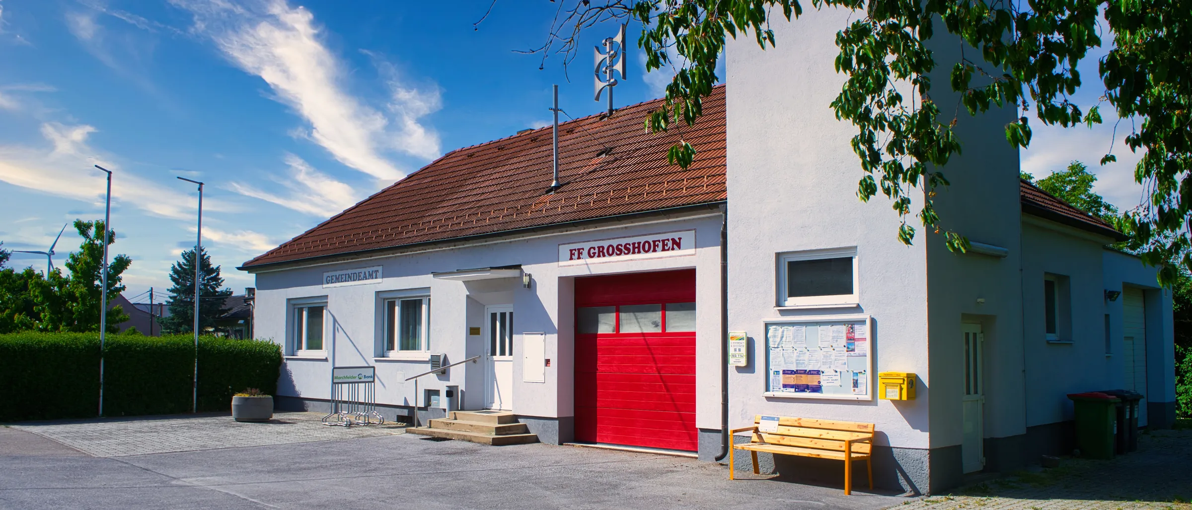 Ein weißes Gebäude mit rotem Dach, roter Garagentür und gelbem Briefkasten. Eine Bank steht neben einem Aushang. Das Gebäude heißt FF Grosshofen.