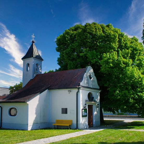 Eine kleine weiße Kirche mit einem braunen Dach und einem Glockenturm steht in einem Grasbereich. Ein gelber Stuhl ist vor der Kirche platziert. Hinter der Kirche befindet sich ein großer Baum.