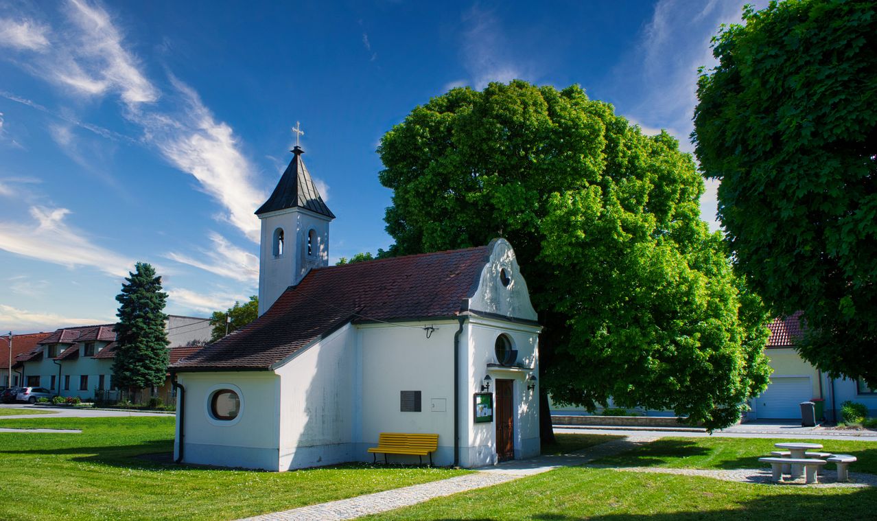 Eine kleine weiße Kirche mit einem braunen Dach und einem Glockenturm steht in einem Grasbereich. Ein gelber Stuhl ist vor der Kirche platziert. Hinter der Kirche befindet sich ein großer Baum.