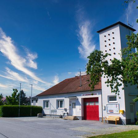 Ein weißes Gebäude mit braunem Dach und roter Garagentür steht unter einem klaren blauen Himmel. Das Gebäude hat mehrere Fenster und einen Turm auf der rechten Seite. Davor steht eine gelbe Bank und ein kleiner Garten.