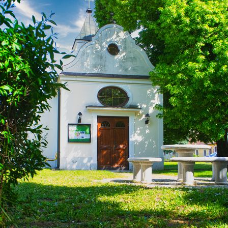 Eine kleine Kapelle mit einer Holztür und einem Kreuz auf der Spitze ist von Grün umgeben. Bänke und ein Tisch sind im Vordergrund.