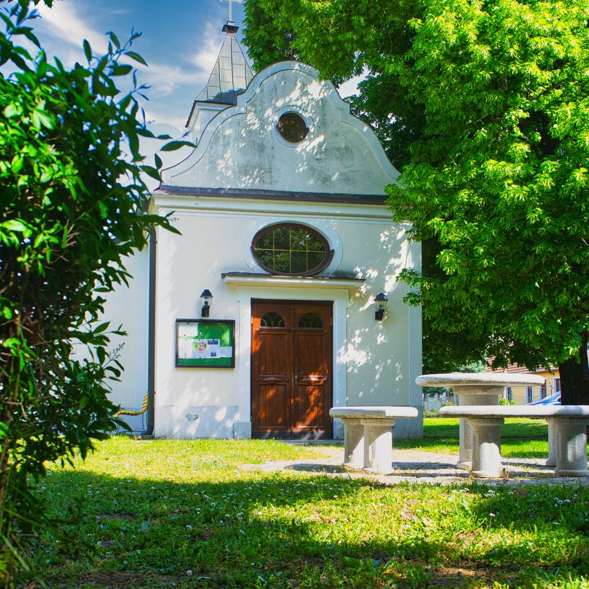 Eine kleine Kapelle mit einer Holztür und einem Kreuz auf der Spitze ist von Grün umgeben. Bänke und ein Tisch sind im Vordergrund.