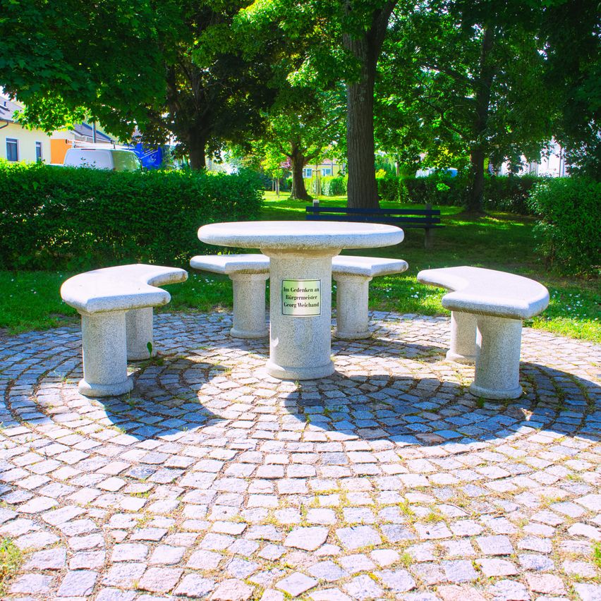 Ein runder Tisch mit Bänken ist von einem Kopfsteinpflasterkreis umgeben, mit einem Schild und einer Parkbank im Hintergrund.