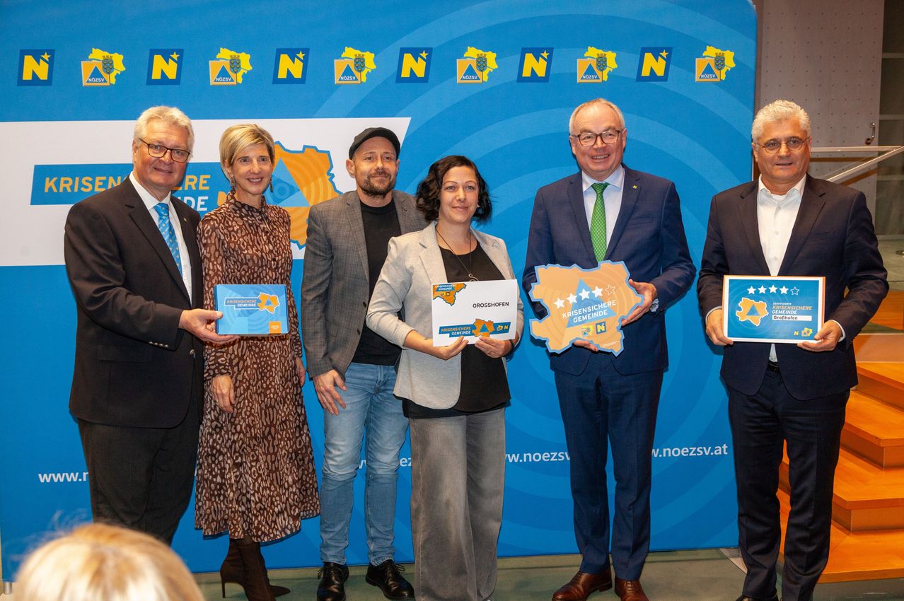 Fünf Personen stehen vor einem blauen Hintergrund mit Logos und einer Karte einer Region. Jede hält ein Schild und sie scheinen etwas bei einer Veranstaltung zu präsentieren.