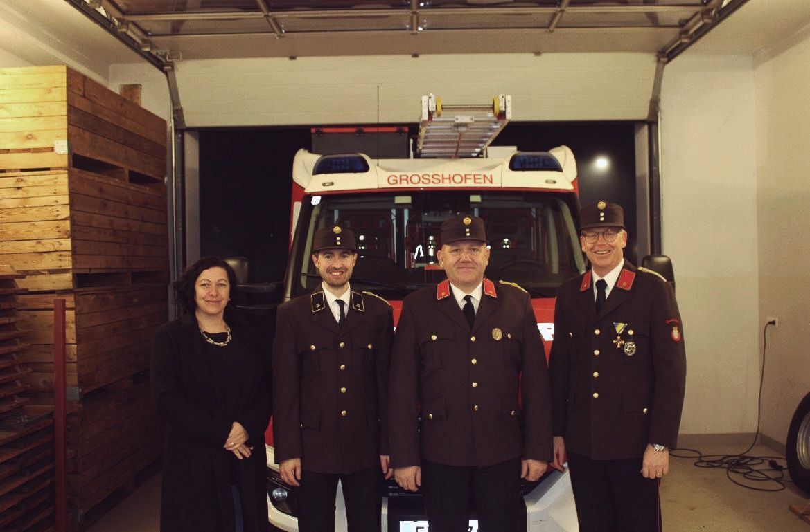 Vier Menschen in Uniform stehen vor einem Feuerwehrauto mit der Aufschrift Grosshofen. Der Mann links lächelt und die Frau links trägt eine Halskette.