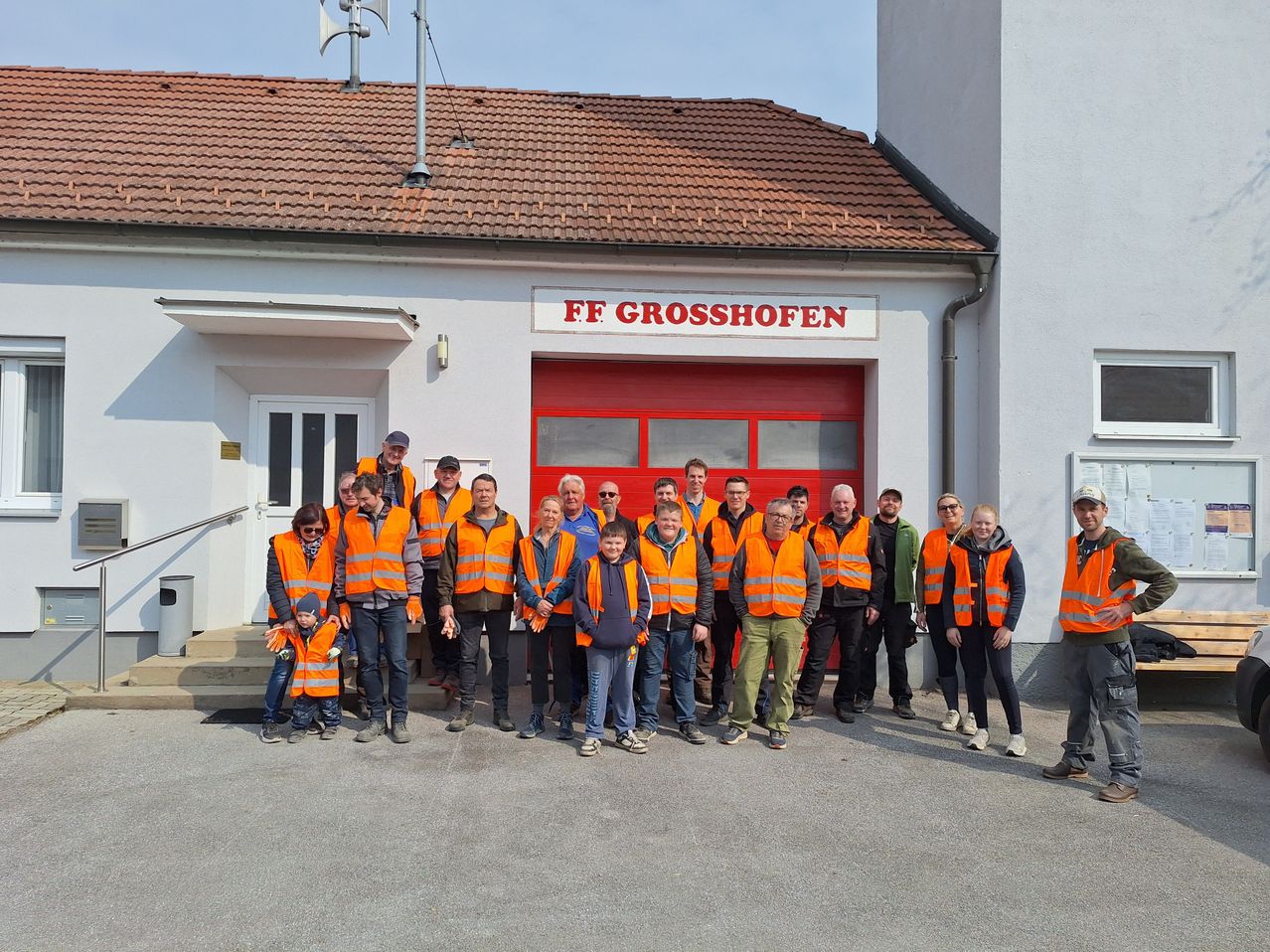 Eine Gruppe von Personen in orange Warnwesten, wahrscheinlich Feuerwehrleute, posiert vor dem FF Grosshofen Feuerwehrhaus.