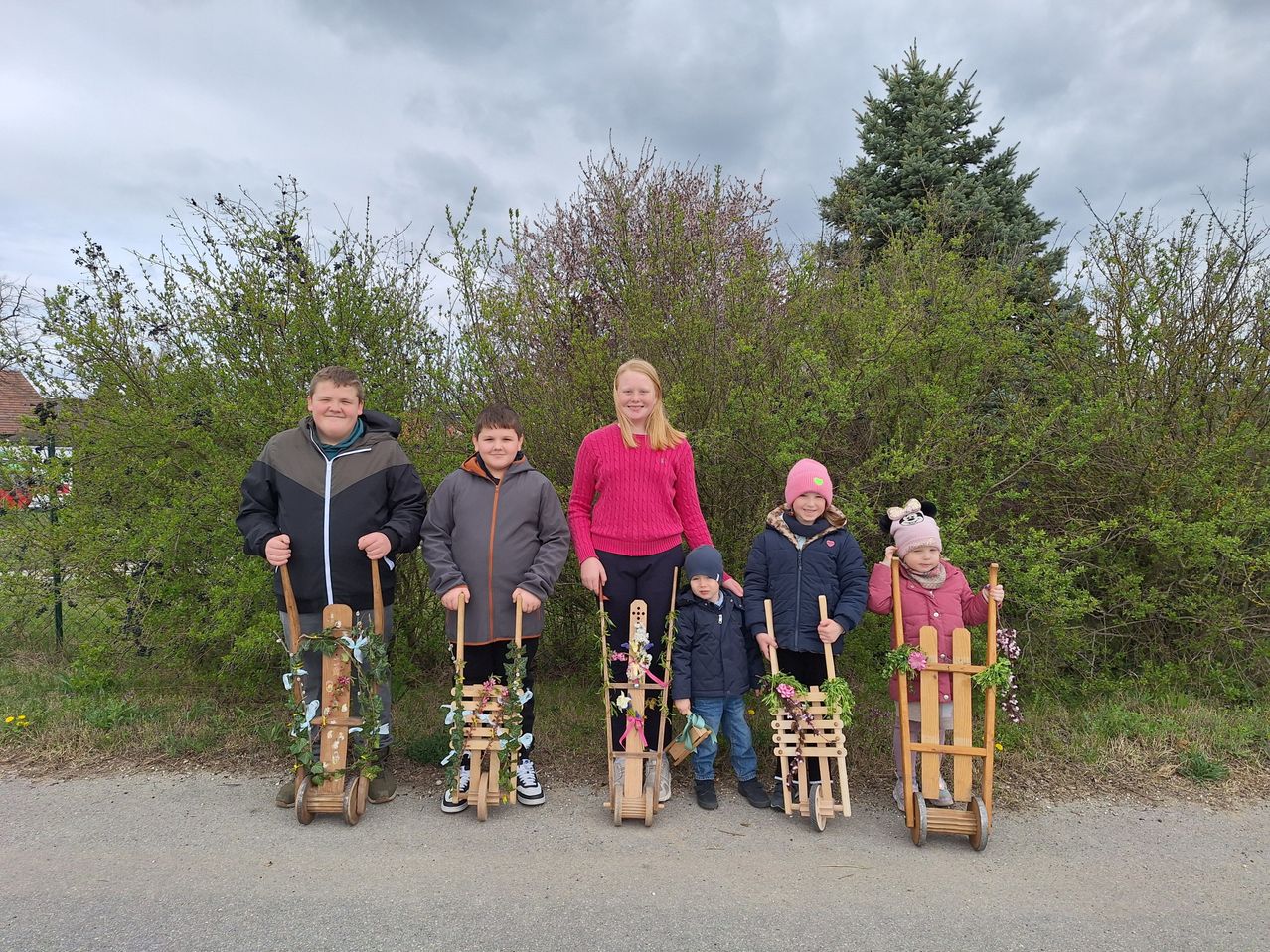 Eine Gruppe von Kindern mit selbstgemachten Schlitten, die mit Blumen und Grün dekoriert sind, posiert für ein Foto auf einem asphaltierten Weg.