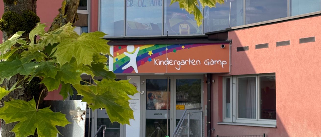 Die Vorderseite eines Kindergartens mit einem bunten Schild, das 'Kindergarten Gampl' lautet. Das Gebäude hat Glaswände und eine Treppe, die zum Eingang führt. Davor steht ein Baum und ein Zaun mit Spielgeräten.