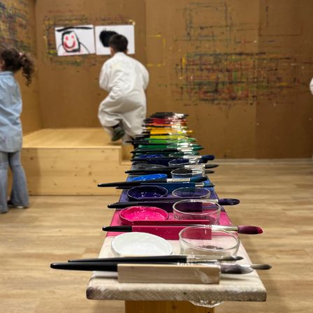 In einem Studio malen Kinder mit Pinsel und Farben. Ein Junge steht auf einer Bank und ein Mädchen steht in der Ecke.