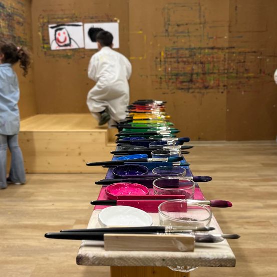 In einem Studio malen Kinder mit Pinsel und Farben. Ein Junge steht auf einer Bank und ein Mädchen steht in der Ecke.