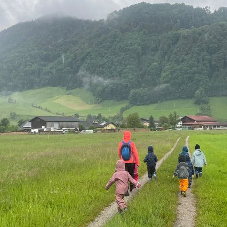 Mehrere Kinder gehen auf einem grasbewachsenen Pfad, tragen Regenmäntel und haben Rucksäcke dabei. Der Pfad wird von saftigem, grünem Gras gesäumt, mit Häusern und einem Berg im Hintergrund.