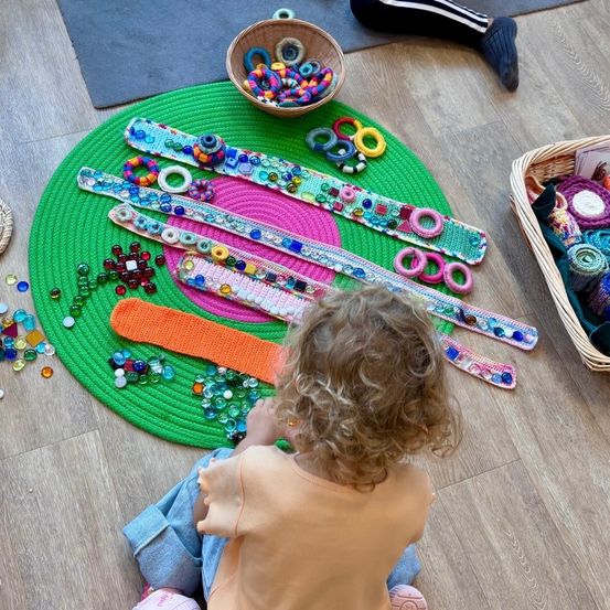 Ein Kind sitzt auf dem Boden, umgeben von bunten gehäkelten Gegenständen, Perlen und Armbändern. Der Boden ist mit einem grünen runden Teppich bedeckt.