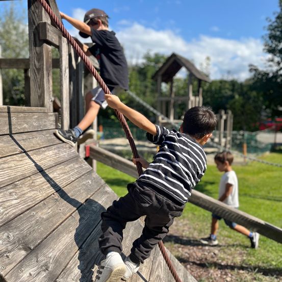 Drei Kinder spielen auf einer Holzspielanlage. Ein Kind klettert an einem Seil, während ein anderes zuschaut. Das dritte Kind läuft auf die Anlage zu. Die Umgebung ist eine Rasenfläche mit Bäumen im Hintergrund.