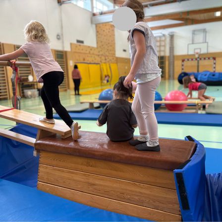 Drei junge Mädchen üben das Gleichgewicht in einer Turnhalle. Ein Mädchen steht auf einem schrägen Brett, während ein anderes darunter sitzt. Dahinter spielt ein Mädchen mit einem Ball.
