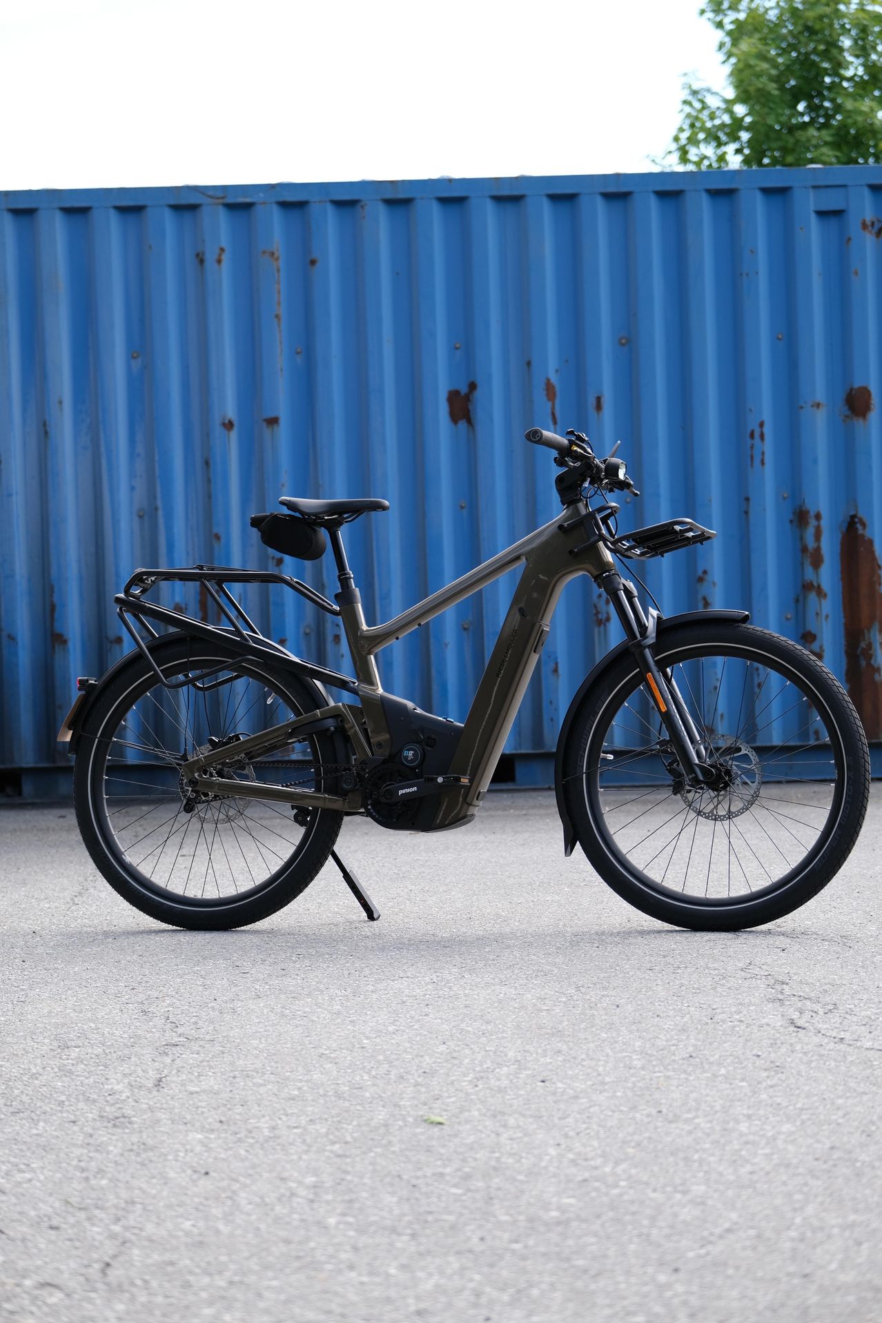 Ein olivgrünes E-Bike mit Heckträger steht vor einer blauen Metallwand mit Rostflecken.