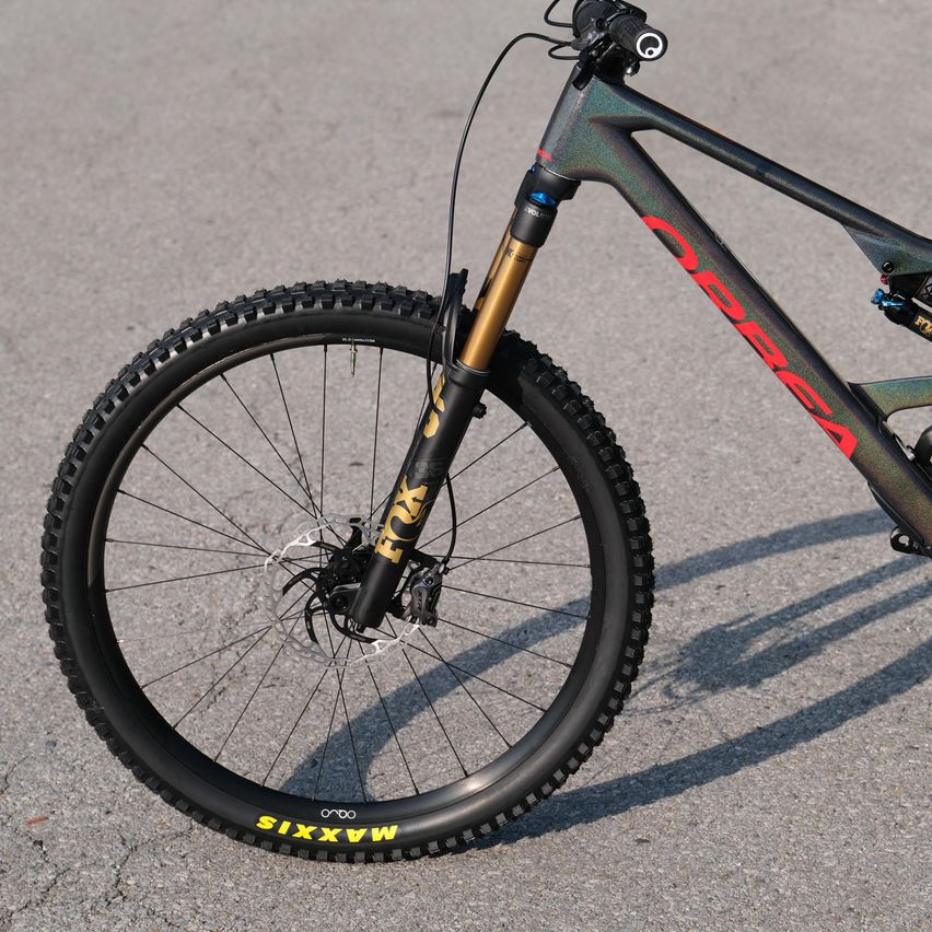 Nahaufnahme des Vorderrads eines Mountainbikes mit schwarzen Reifen, silbernen Speichen und einem gelben FOX-Logo am Gabel. Der Rahmen ist grün mit rotem Text, der OBSIDIAN liest.