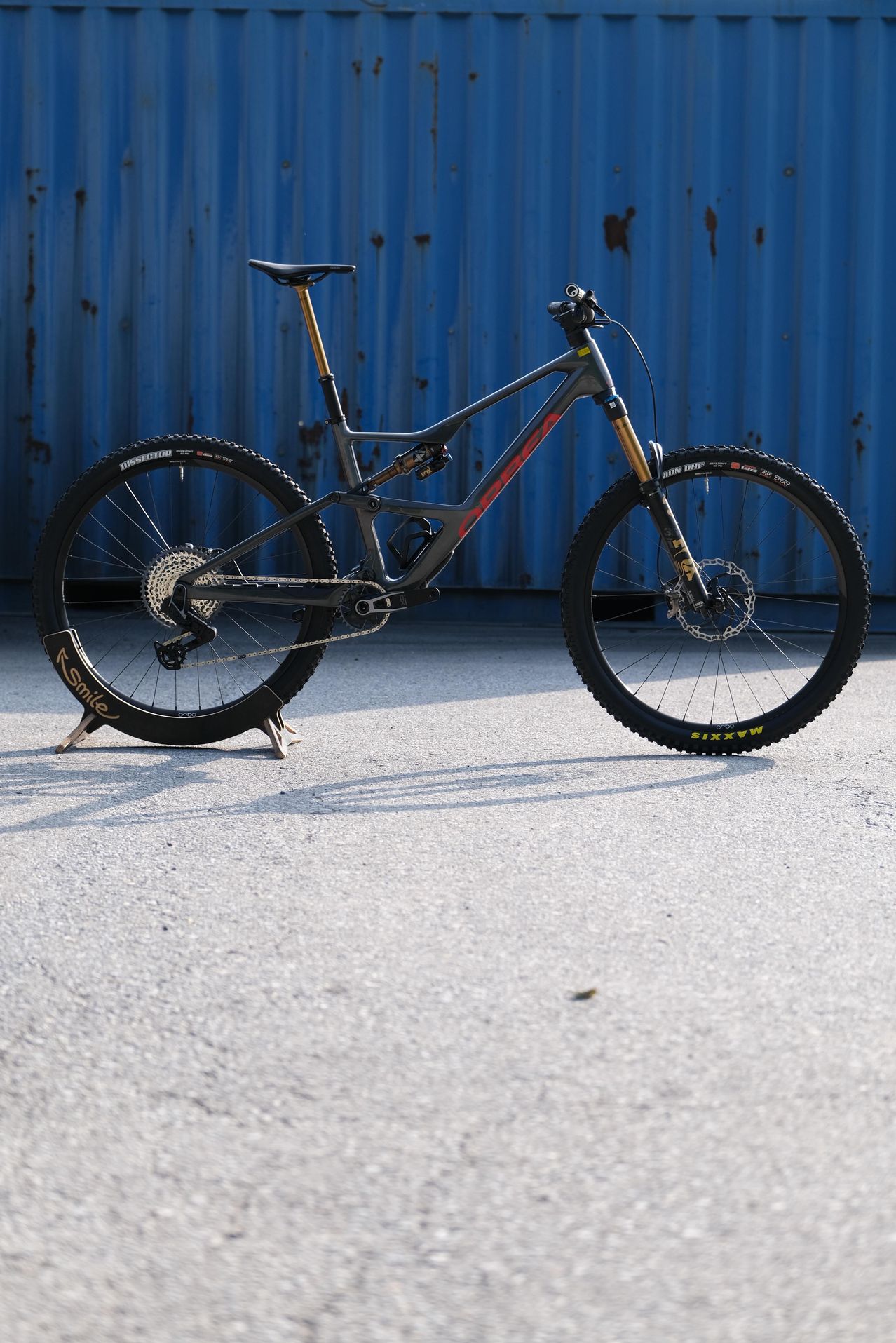Ein schwarzes Mountainbike steht vor einer blauen Metallwand. Es hat einen schwarzen Rahmen, rote Akzente und einen schwarzen Sattel. Die Vorder- und Hinterreifen haben gelbe Schriftzüge.