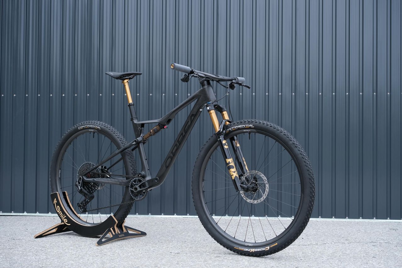 Ein schwarzes Mountainbike mit goldenen Akzenten ist vor einer grauen Metallwand ausgestellt. Das Fahrrad verfügt über breite Reifen und ein Federungssystem, geeignet für Offroad-Gelände.