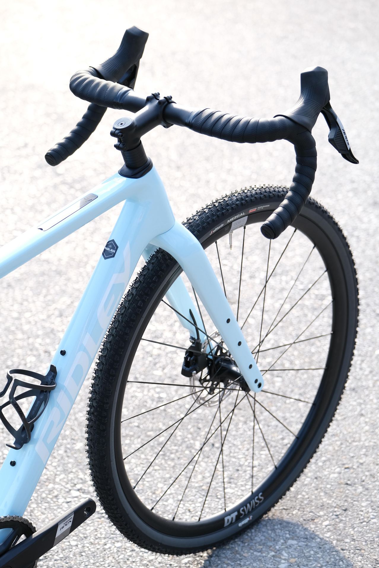 Nahaufnahme eines weißen Fahrrads mit schwarzen Reifen und Lenker auf einer asphaltierten Oberfläche. Das Fahrrad hat einen Flaschenhalter am Rahmen.