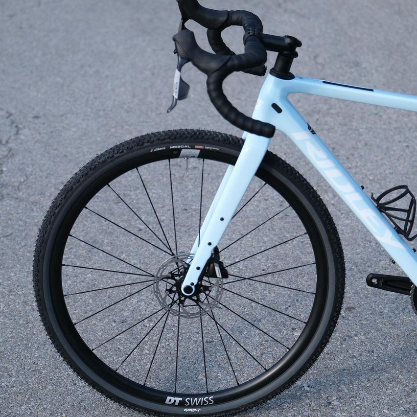 Nahaufnahme des vorderen Rads eines weißen Fahrrads mit schwarzen Reifen und silbernen Speichen, geparkt auf einer Betonoberfläche.