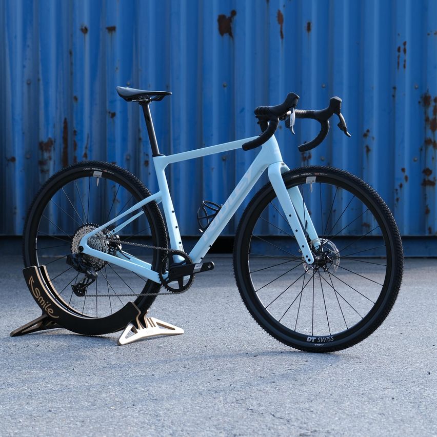 Ein weißes Rennrad mit schwarzen Felgen und Reifen wird auf einer Betonfläche mit einer blauen Metallwand im Hintergrund ausgestellt.