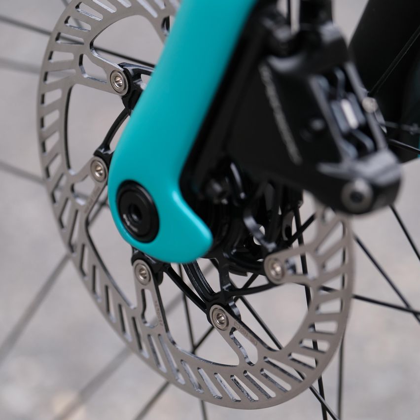 Eine Nahaufnahme des Vorderrads eines Fahrrads zeigt einen türkisen Metallrahmen und eine silberne Bremsscheibe mit schwarzen Bremssätteln.