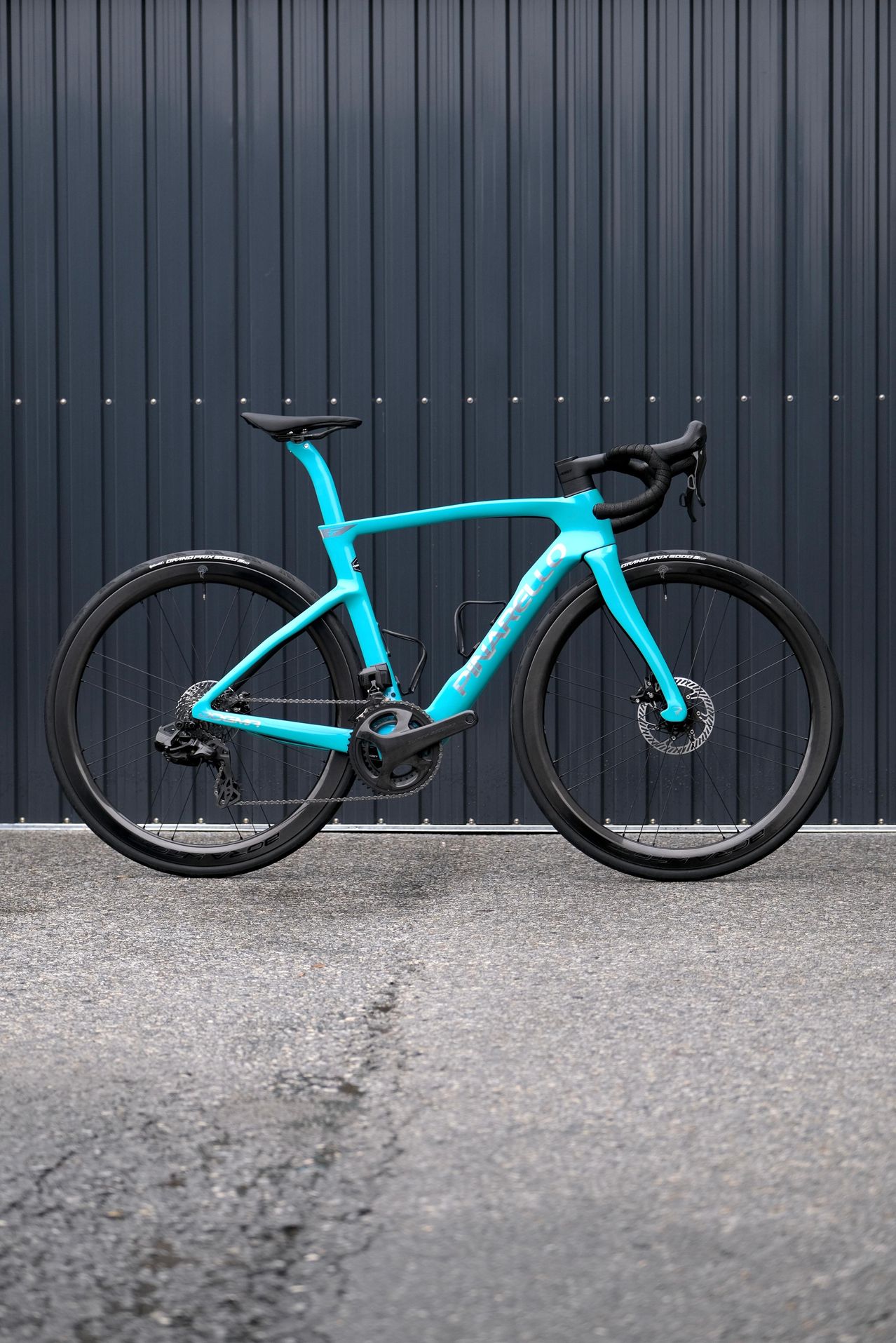 Ein türkisfarbenes Rennrad mit schwarzen Lenkern und Rädern steht vor einer grauen Metallwand.