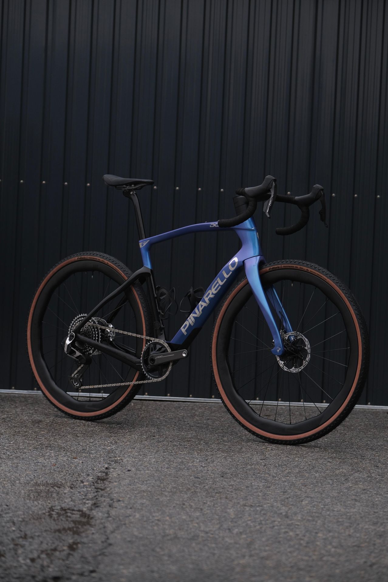 Ein blaues Pinarello-Fahrrad mit orangefarbenen Reifen steht vor einer schwarzen Metallwand.