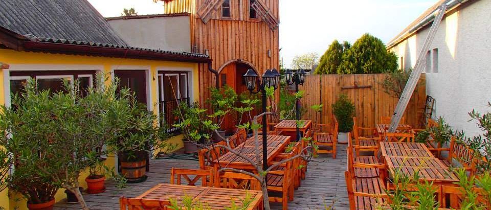 Ein hölzerner Außenbereich mit Tischen und Stühlen unter einer Windmühle. Ein gelbes Haus mit Fenstern und einem Schornstein ist in der Nähe. Bäume und ein Holzzaun umgeben den Bereich.