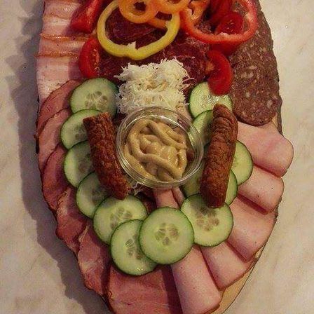 Ein großer Holzteller zeigt verschiedene Fleischsorten, Gurken, geschnittene Paprika, Käse und eine Schüssel mit Senf auf einer Marmoroberfläche.