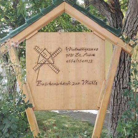 Ein hölzernes Schild mit einem Windmühlenbild befindet sich vor einem Gebäude mit gelber Wand und grüner Umrandung. Das Schild trägt die Aufschrift 'Windmuehlgasse 40, 7161 St. Andrae, 0066416820153, Buschenschank zur Mühle.' Das Schild ist von grünem Gras und rosa Blumen umgeben.