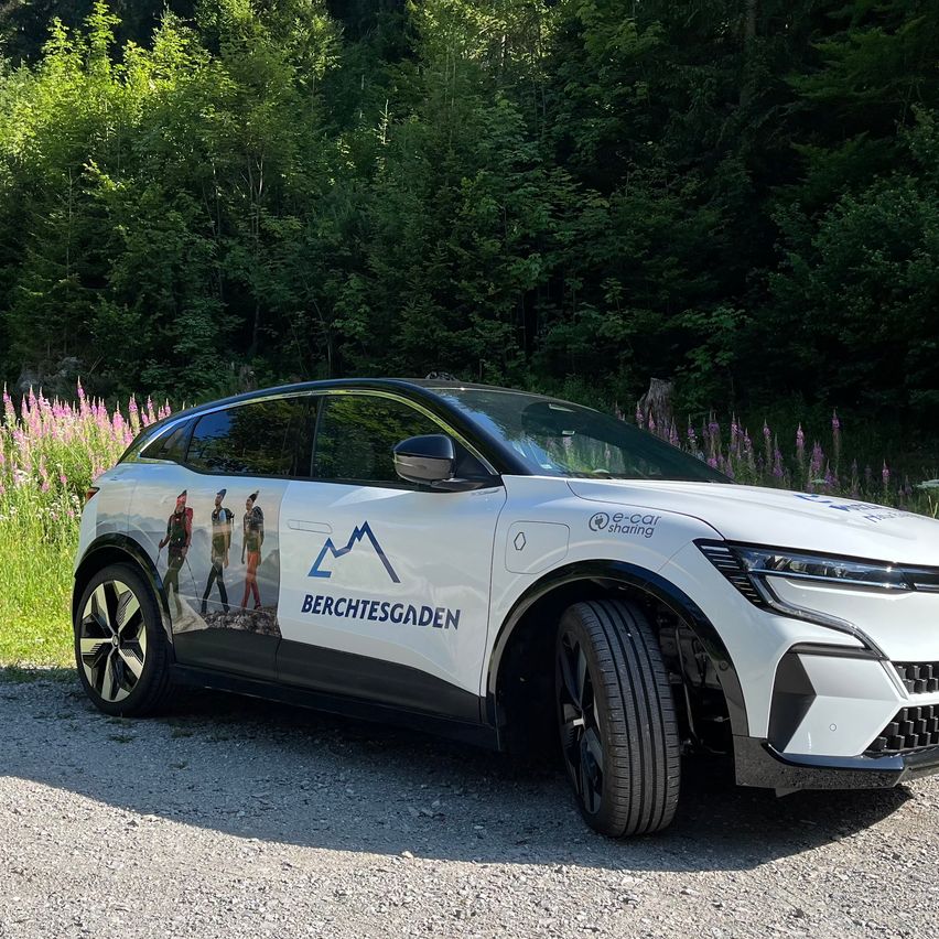 Ein weißer Elektrowagen mit Logo und dem Wort 'Berchtesgaden' auf einem Kiesweg mit Hintergrund aus grünen Bäumen und Blumen.