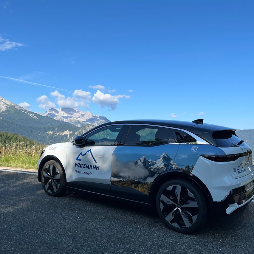 Ein weißer Elektrowagen mit blauem Berglogo steht auf einer Straße mit einer malerischen Bergkette im Hintergrund.