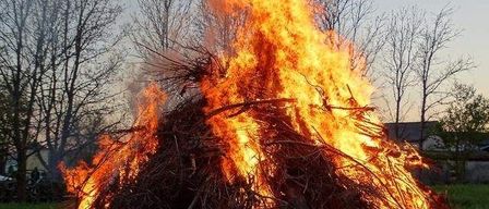 Ein großes Lagerfeuer brennt hell in einem offenen Feld, umgeben von Bäumen. Das Feuer besteht aus Ästen und Zweigen.