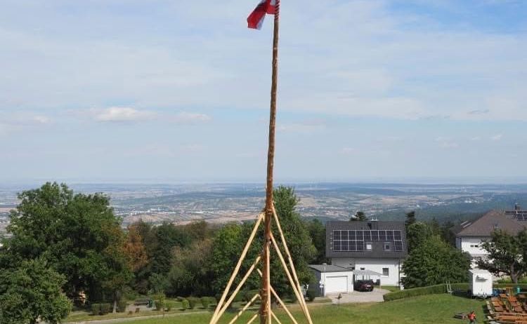 Eine Gruppe von Menschen umgibt einen hohen Holzpfahl mit einer Flagge auf einem Hügel unter blauem Himmel.