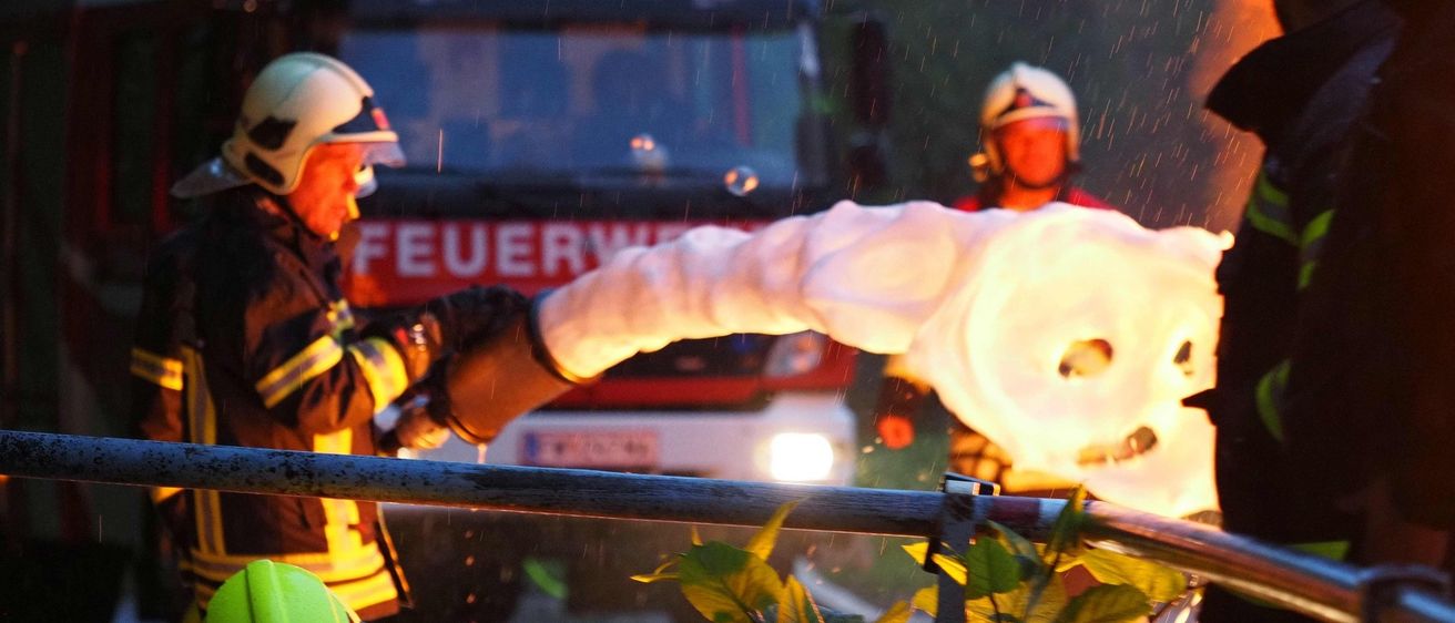 Feuerwehrleute mit weißen Helmen und Feuerwehranzügen sprühen Wasser im Regen. Ein Feuerwehrwagen ist hinter ihnen geparkt, und ein anderer Feuerwehrmann hält eine Schlauch.