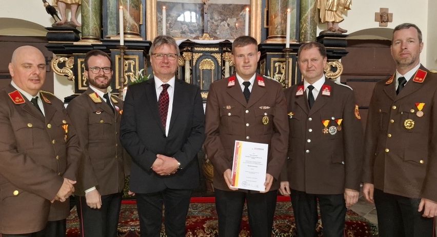 Fünf Männer in Militäruniformen stehen zusammen vor einem Altar. Der Mann in der Mitte hält ein Zertifikat und lächelt.