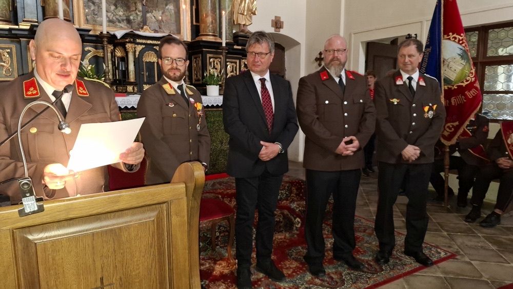Fünf Männer in Militäruniformen und Anzügen stehen in einer Kirche, einer Mann hält ein Stück Papier und ein anderer Mann spricht in ein Mikrofon.